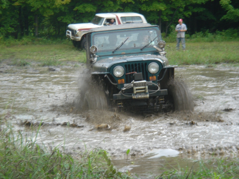 Green Jeep CJ-7 driving through a mud puddle, white Ford Bronco behind it
