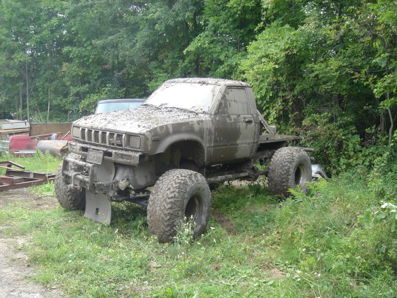 Mud-covered pickup truck with large tires parked in grass near trees. Another vehicle is partially visible behind it