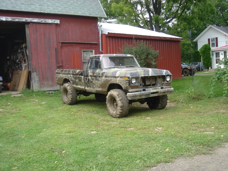 Muddy, camouflage-painted pickup truck parked on grass in front of a red barn