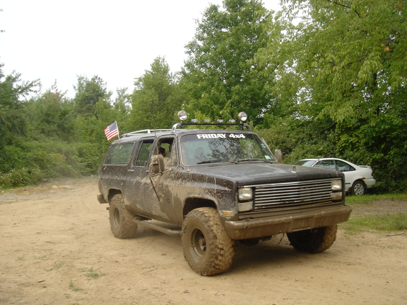 Muddy brown SUV with off-road tires, roof rack, and American flag. Text on windshield reads 'FRIDAY 4X4'