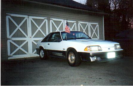 1989 Ford Mustang - This 1989 Ford Mustang GT is captured in a three-quarter front view, parked outside a garage with an American flag on its antenna.