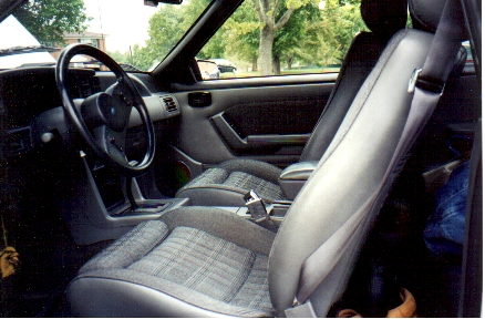 1989 Ford Mustang - The interior of a car shows plaid fabric seats and a dashboard with a black steering wheel.