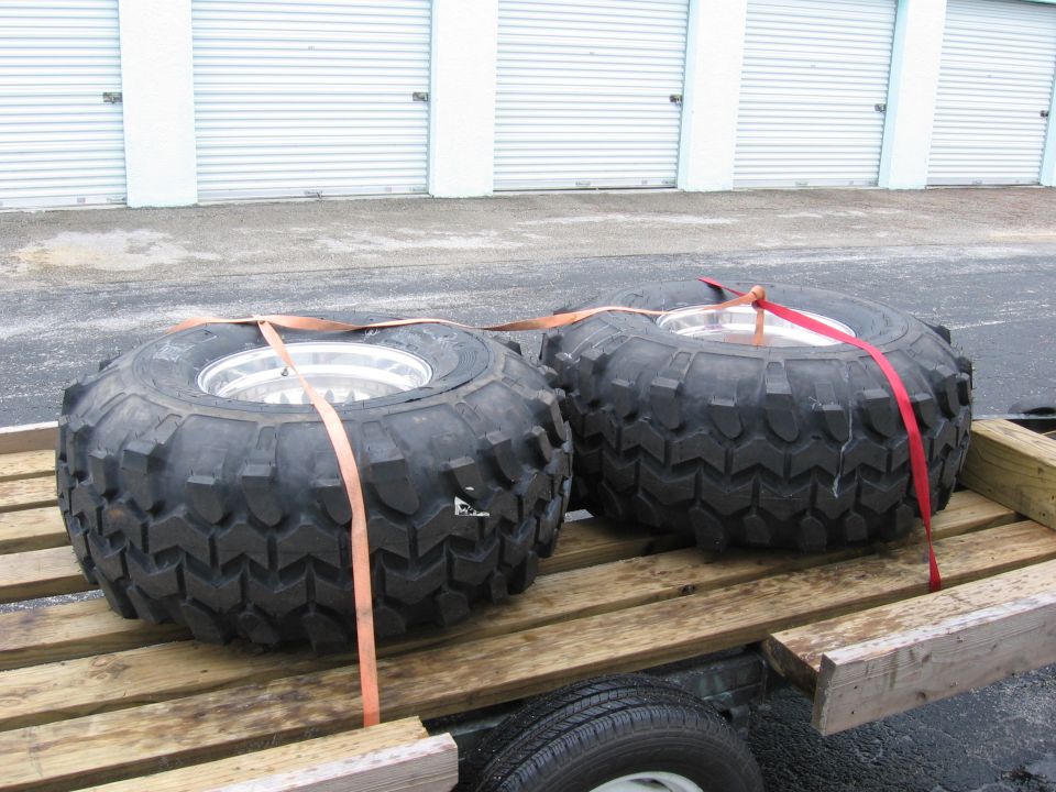 Two large off-road tires with aggressive tread secured with straps on a wooden trailer bed