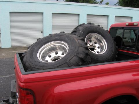 1974 Ford F-100 - Two oversized tires with chrome rims are transported in the bed of a red pickup truck.