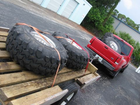 1974 Ford F-100 - Oversized tires are strapped to a wooden trailer, with a red Ford Ranger pickup truck visible behind it.