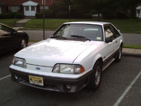 1989 Ford Mustang - A white 1989 Ford Mustang GT is parked in a lot, showing its front 3/4 view and license plate.
