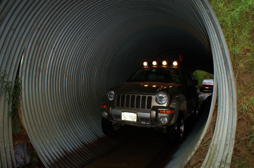 Jeep Liberty with light bar driving through a large metal culvert. Another red vehicle is visible further down the tunnel