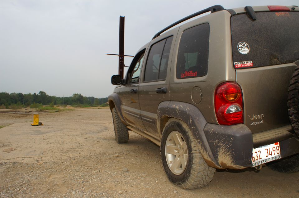 Rear view of a tan Jeep Liberty parked on a dirt lot. It has mud on the fenders and a spare tire mounted on the back