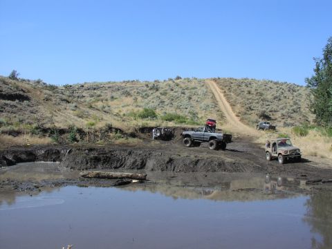 1979 Ford Bronco - Several off-road vehicles are seen navigating a challenging muddy terrain under a clear blue sky, with a dirt track ascending a distant hil…