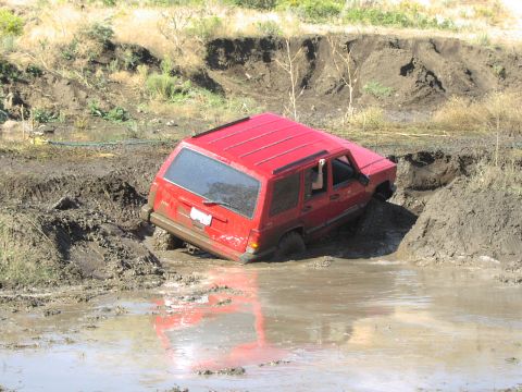1979 Ford Bronco - A red SUV is deeply stuck in a muddy ditch, with its front wheels submerged in water.
