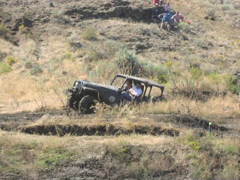 1979 Ford Bronco - A black Jeep Wrangler is shown traversing a challenging muddy course with spectators watching from a hillside.