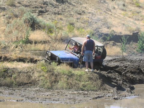 1979 Ford Bronco - An off-road vehicle navigates a muddy terrain, with two people inside and another person observing.