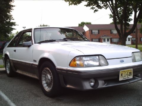 1989 Ford Mustang - A white 1989 Ford Mustang GT is parked in a lot with buildings and trees in the background.