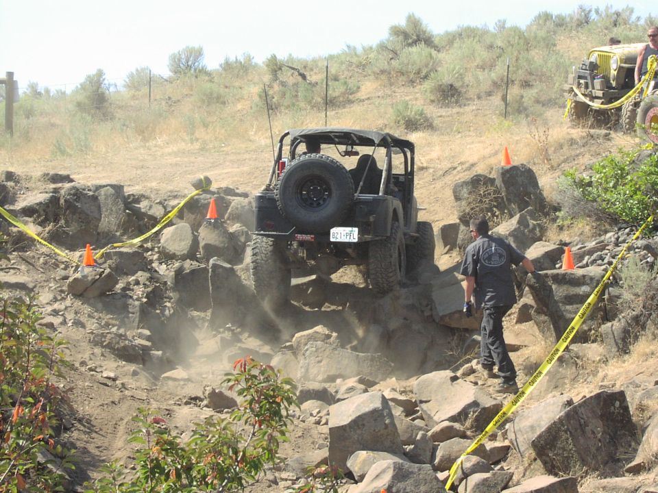 Black Jeep Wrangler navigating a rocky off-road course, rear view, with a yellow caution tape barrier