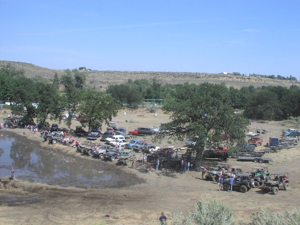 Many off-road vehicles parked on dirt and near a muddy pond under a clear blue sky