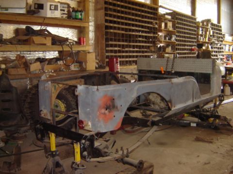 1992 Ford Bronco - The chassis of a Jeep CJ-6 is shown in a workshop, supported by jack stands, with its body panels partially disassembled.