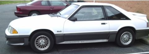 1989 Ford Mustang - A white coupe with dark gray trim and a rear spoiler is parked next to a maroon sedan.