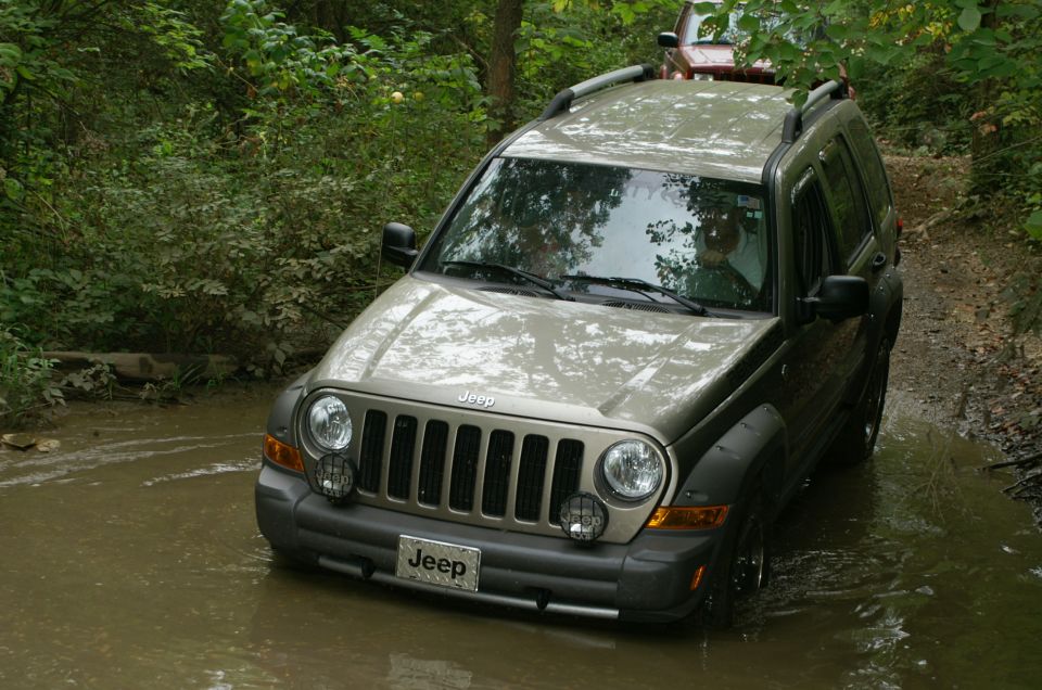 2006 Jeep Liberty driving through a muddy, wooded area; another vehicle is visible behind it