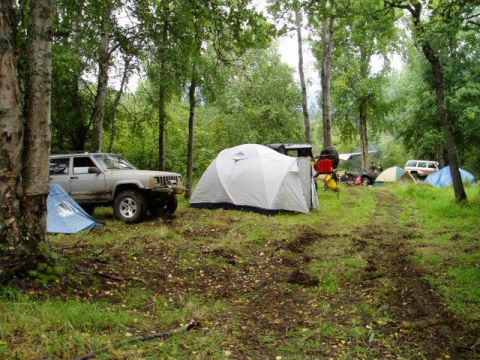 2000 Ford E350 - A silver Jeep Cherokee XJ is parked at a campsite, surrounded by tents and trees. Another vehicle is visible in the background.