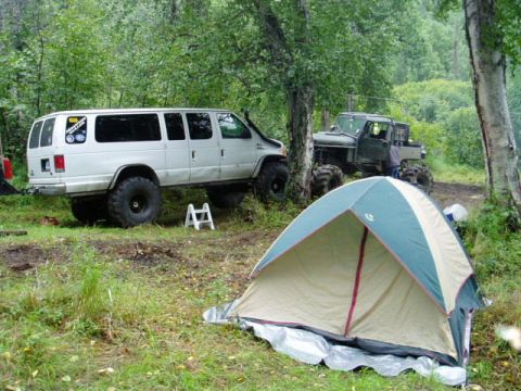 2000 Ford E350 - A white van and an off-road truck are parked in a forest clearing with a tent set up in the foreground.