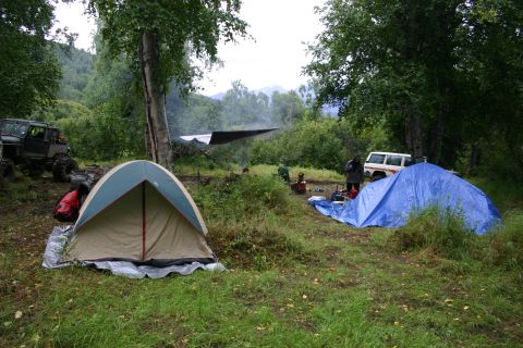 2003 Jeep TJ - A campsite is set up in a wooded area with two tents, a blue tarp, and two vehicles.