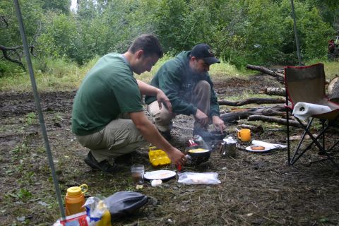 2003 Jeep TJ - Two men prepare food over a campfire at a campsite, with cooking supplies and camping chairs visible.