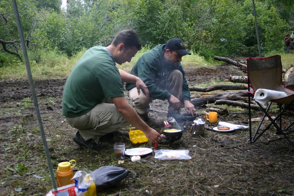 Two campers cooking eggs in a skillet over a camp fire with percolator and plates on muddy ground