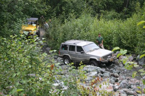 2003 Jeep TJ - A silver Jeep Cherokee XJ navigates a rocky trail with a yellow Jeep in the background.
