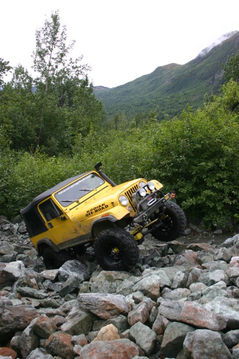 2003 Jeep TJ - A yellow Jeep navigates a rocky terrain with lush green foliage and mountains in the distance.
