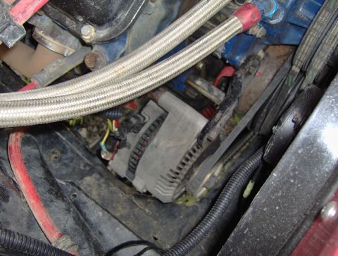 1978 Ford Bronco - Engine-bay detail of a cast-aluminum alternator mounted low on a blue engine block, with its plug-in wiring harness and V-belt visible. Two…