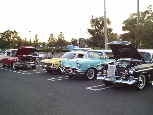 Several classic cars parked in a lot, hoods up on some. One has a '1955 NOMAD' badge