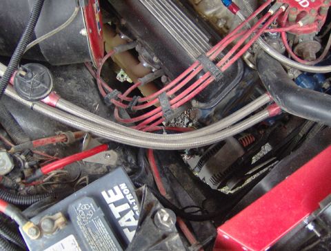 1978 Ford Bronco - Cluttered engine bay looking down on a black finned valve cover with red MSD plug wires in looms, cast exhaust manifold, and braided stainl…