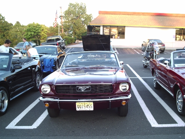 Maroon 1965 Ford Mustang convertible parked in a lot, with another Mustang and a motorcycle nearby