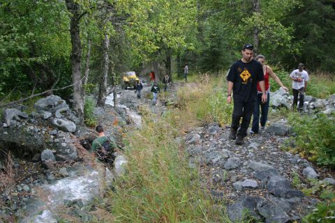 2003 Jeep TJ - A group of people navigate a rocky trail alongside a stream, with a yellow off-road vehicle visible in the background.