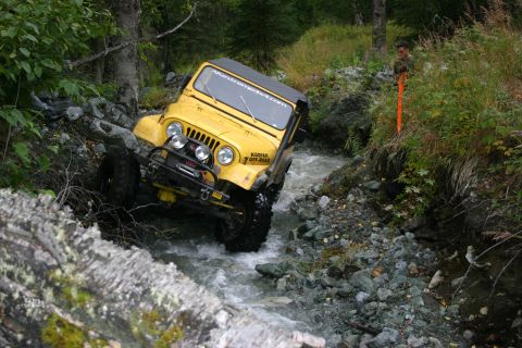 2003 Jeep TJ - A yellow Jeep CJ navigates a rocky stream, its tires kicking up water as it moves through the off-road terrain.