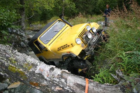 2003 Jeep TJ - A yellow Jeep CJ-7 navigates a difficult off-road course, crossing over a fallen tree in a stream.