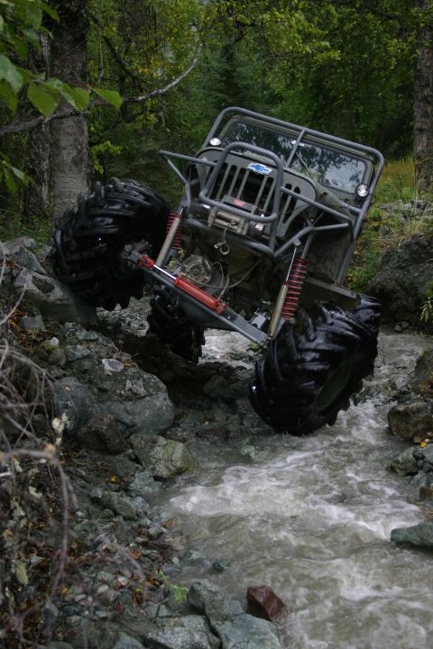 2003 Jeep TJ - An off-road vehicle with oversized tires drives through a rocky stream in a wooded area.