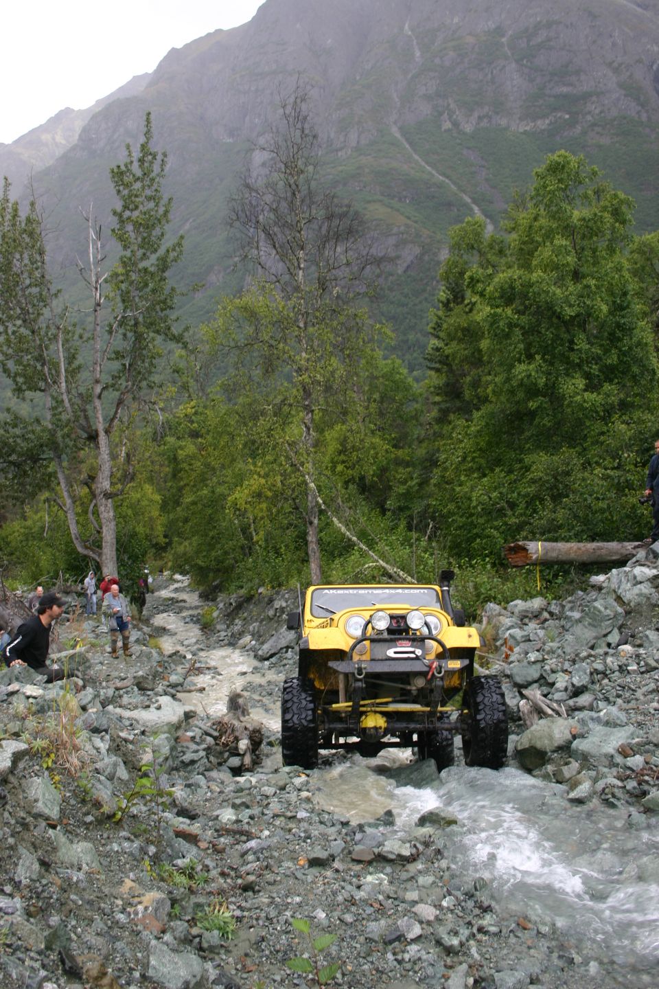 Yellow Jeep Wrangler driving through a rocky stream with people watching