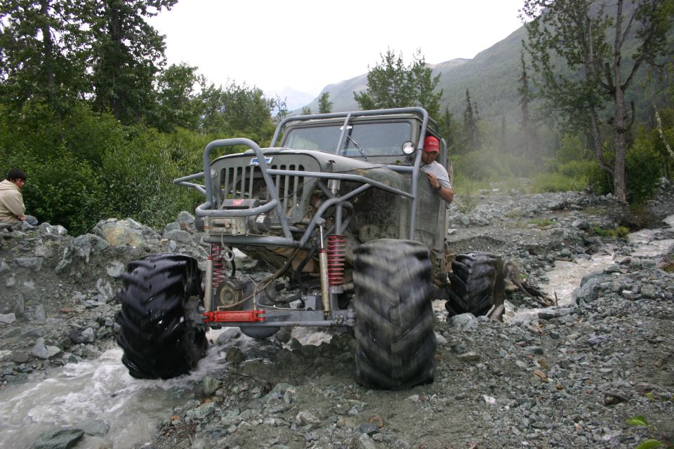 Modified Jeep CJ-7 with large tires driving through a rocky stream bed, person in red hat visible in driver's seat