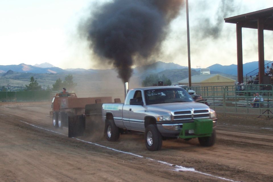 Silver Dodge Ram pickup truck with large exhaust stack emitting black smoke, pulling a sled in a competition. Green front bumper/weight