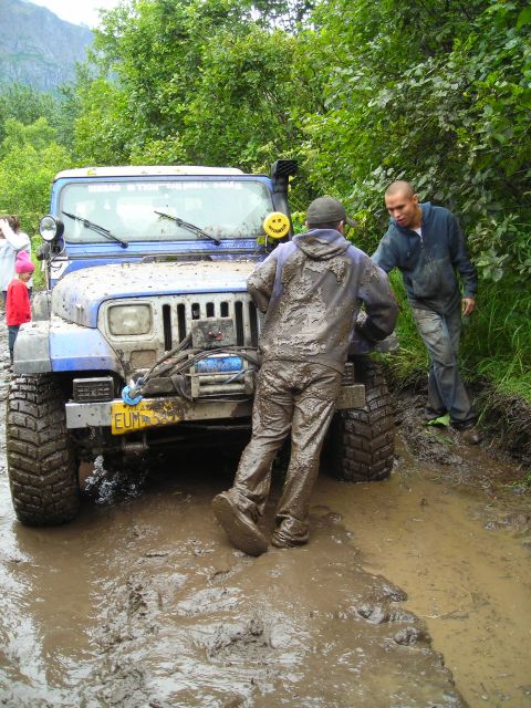 2003 Jeep TJ - A blue 2003 Jeep TJ is stuck in deep mud, with two people attempting to push it free.