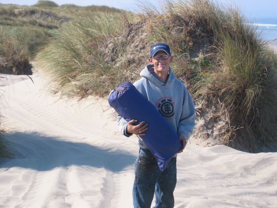 Man in hoodie and jeans holds blue sleeping bag on sandy dunes near ocean