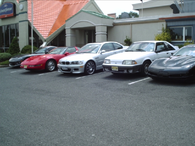 Several sports cars, including a white Ford Mustang, a red Chevrolet Corvette, and a silver BMW, are parked in a lot outside a building