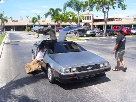 1994 Ford Probe - A silver DMC car with gullwing doors open is being loaded with a box by a person inside, while another person walks nearby.