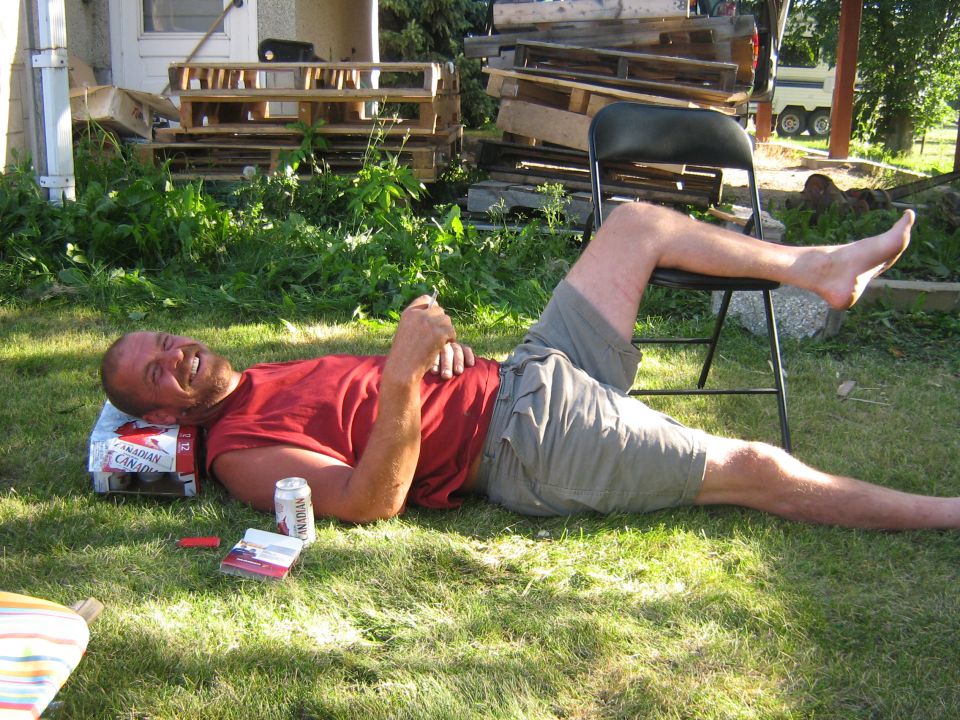 Man in red shirt and shorts lying on grass, propped on a box, with a chair and pallets in background