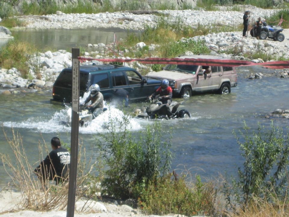 Two ATVs splash through a river, with a dark green SUV and a tan Jeep Cherokee behind them