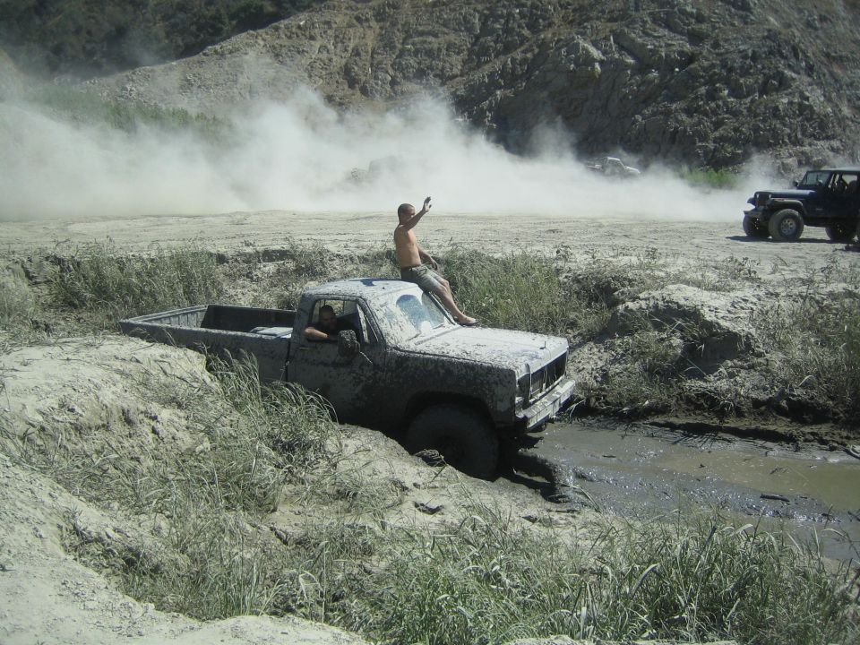 Guy sitting on roof of muddy pickup stuck in mud pit. Another guy in cab. Dust cloud in background with other off-road vehicles