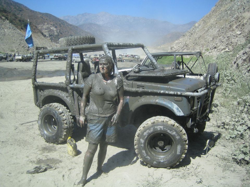 Mud-covered woman standing next to a mud-covered 1987 Suzuki Samurai. Several other off-road vehicles are visible in the background