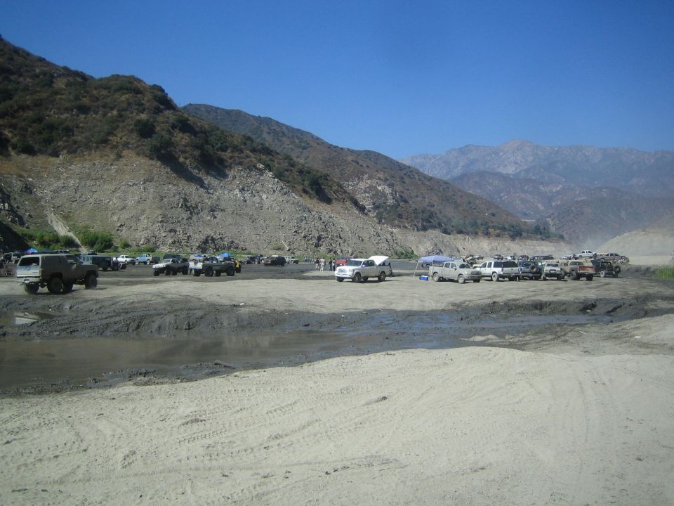 many SUVs and trucks parked in a sandy, rocky area with mountains in the background