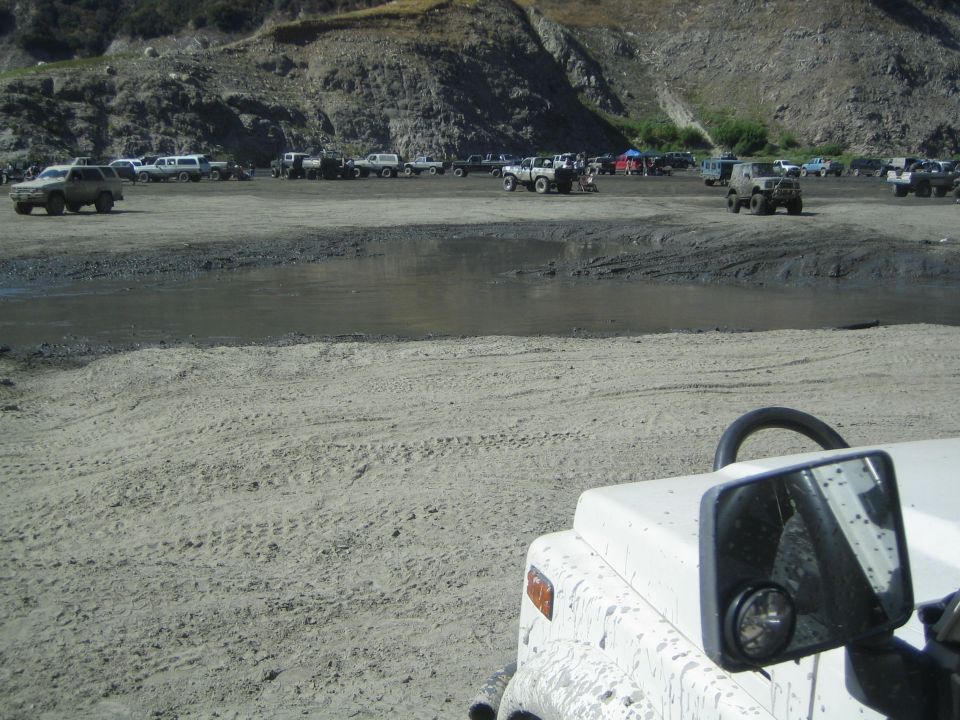 many off-road vehicles parked in a dirt and mud lot near a small body of water, with a rocky hillside in the background